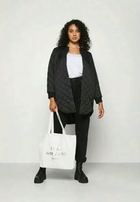 Veste matelassée noire avec une coupe ample, portée sur une chemise blanche et un pantalon noir. Le modèle tient un sac fourre-tout blanc avec un texte argenté. Bottes noires à lacets.