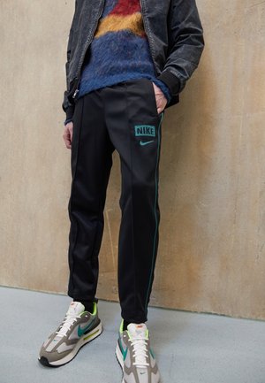Person iført sorte Nike træningsbukser med turkis logo og piping, farverig fluffy sweater, mørk jakke og beige sneakers med blågrønne detaljer.
