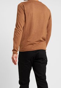 Chemise marron à manches longues en matière douce, avec un col rond et une coupe ajustée, associée à un jean noir.