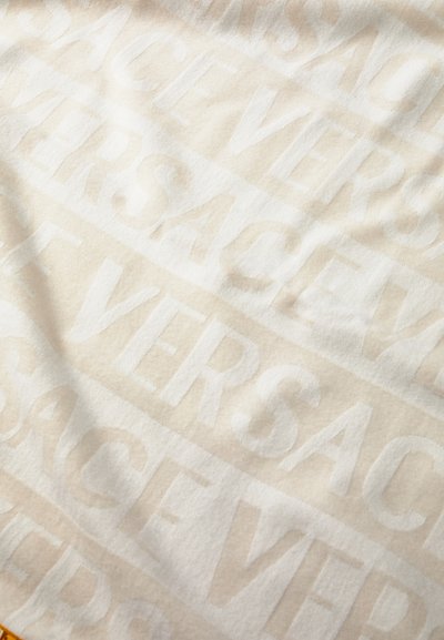 Bijela plišana deka s reljefnim uzorkom i riječju "VERSACE" u raznim veličinama. Mekana tekstura, minimalistički dizajn, suptilni sjaj.