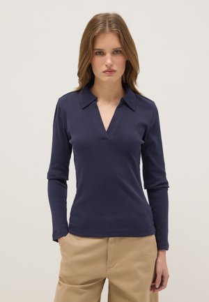 Vrouw met schouderlang haar die een marineblauw poloshirt met lange mouwen en beige broek draagt, staand tegen een effen lichte achtergrond.