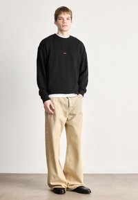 Μαύρο βαμβακερό φούτερ με κόκκινο λογότυπο, φορεμένο πάνω από άσπρο πουκάμισο. Συνδυασμένο με αμυγδαλί τζιν με φαρδιά γραμμή και μαύρα παπούτσια.