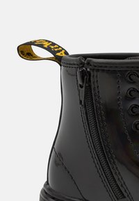 Stivale alla caviglia in vernice nera con zip laterale, punta rotonda e un pratico anello con logo giallo. Texture liscia con dettagli lacci.