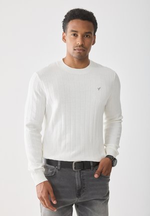 Suéter blanco de canalé con cuello redondo, mangas largas y un sutil patrón texturizado, combinado con jeans de mezclilla grises y un cinturón negro.
