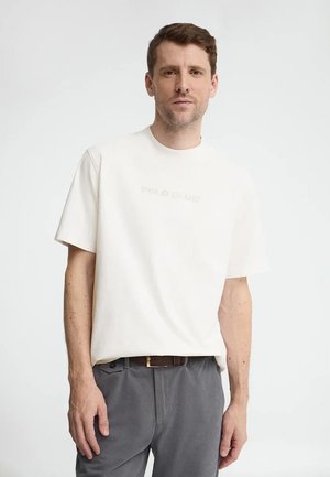 Mężczyzna z krótkimi brązowymi włosami, ubrany w zwykły biały t-shirt i szare spodnie, stoi na jednolitym jasnym tle.