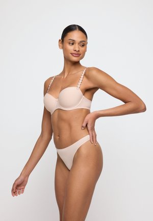 Vrouw poseert in een licht beige lingerieset met geschulpte bandjes tegen een effen witte achtergrond.