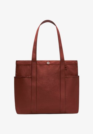 Bolso tote grande de color marrón rojizo con dos asas largas, textura suave, bolsillos laterales y un pequeño logo circular en la parte superior central.