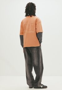 Kortærmet orange t-shirt med grafiktryk på ryggen, parret med løst siddende mørkegrå jeans og sorte sneakers.