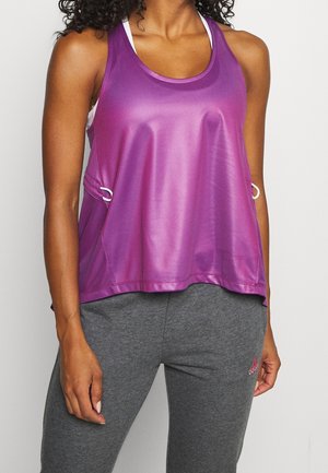 Femme portant un haut de sport sans manches violet brillant et un pantalon de survêtement gris Adidas, debout avec une main détendue le long du corps.