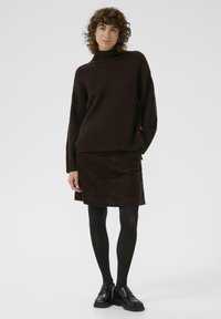 Pull en col roulé marron avec un ourlet côtelé, associé à une jupe en velours côtelé marron. Porté avec des collants noirs et des chaussures. Design simple et texturé.