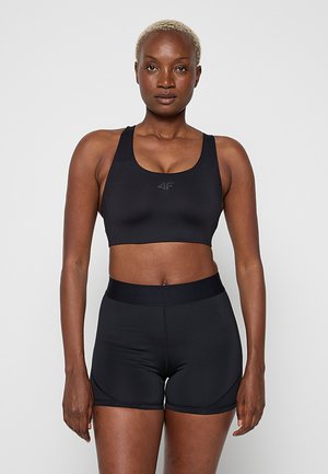 Vrouw met kort blond haar die een zwarte sportbeha en bijpassende strakke sportshorts draagt, staand tegen een effen achtergrond.