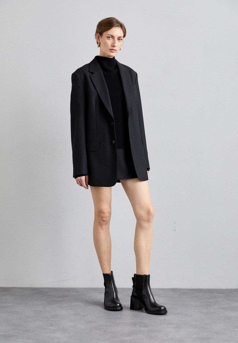 Blazer negro oversize sobre un suéter de cuello cisne negro ajustado, combinado con una minifalda negra y botas de tobillo negras con tacón. Texturas suaves.
