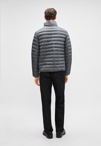 Pelēks pūkains mētelis ar horizontālu quilted dizainu, augstu apkakli un gludu faktūru, apvienots ar tumšiem biksēm un apaviem, parādīts no aizmugures.