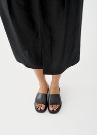Zwarte leren sandalen met brede banden en een platte zool, waarbij de tenen zichtbaar zijn. Het omliggende kledingstuk is een lange zwarte rok. Witte achtergrond.