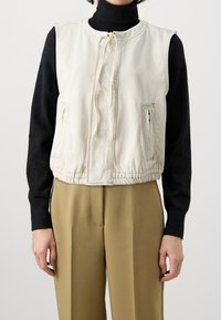 Vesta z lehké béžové látky s kulatým výstřihem, zapínáním na přední zip, dvěma šikmými kapsami a žebrovaným spodním lemem, vrstvená přes černý rolák.