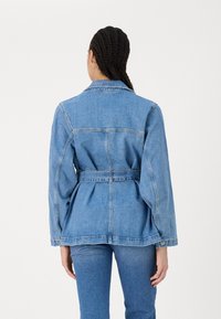 Džínová bunda v světle modré barvě, s páskovým pasem, dlouhými rukávy a standardním límcem. Zadní design zahrnuje švy a peplum efekt.