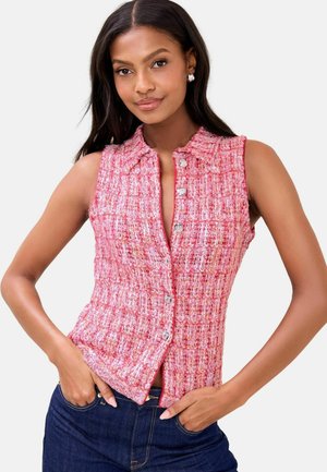 Žena v ružovom bezrukávovom textúrovanom blúzovom topu na zapínanie a tmavomodrých džínsach pózuje s rukami v vreckách na bielom pozadí.