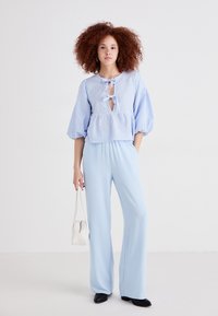 Μπλούζα από μπλε καρό ύφασμα με φουσκωτά μανίκια και κορδόνι στον λαιμό, σε συνδυασμό με ανοιχτό μπλε παντελόνι σε φαρδιά γραμμή και λευκή τσάντα. Μαύρα παπούτσια με τακούνι.