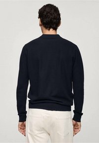 Marineblauer Strickpullover mit einfarbiger Textur, ausgestattet mit einem dezenten gerippten Kragen und Bündchen, kombiniert mit weißen Hosen.