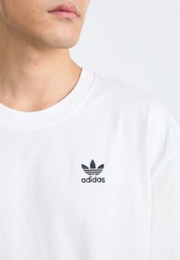 Tricou alb din bumbac, cu un mic logo negru Adidas pe pieptul stâng. Guler rotund și mâneci scurte, textură netedă.