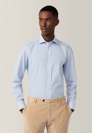 Camisa de botones azul claro con un patrón sutil, de manga larga y con un cuello clásico. Combinada con pantalones de algodón beige.