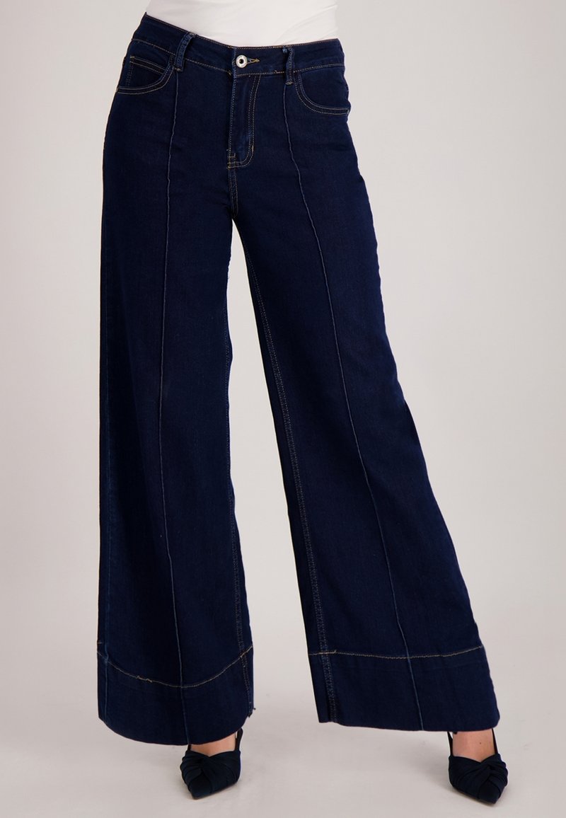 Denimbroek met wijde pijpen in donkerblauw. Voorzien van voorzakken, contrasterende stiknaden en een hoge taille met knoopsluiting.