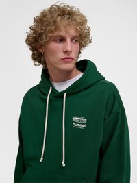 Ung mand med krøllet blondt hår iført grøn Hummel sportshættetrøje, kigger til siden mod en ensfarvet baggrund.