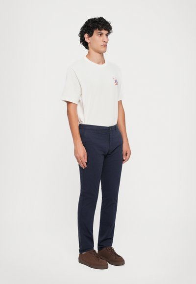 Uomo con capelli ricci in piedi, indossa una maglietta bianca con una piccola grafica colorata, pantaloni blu scuro e sneakers marroni su uno sfondo bianco.