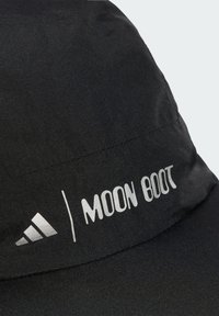 Črna kapa z mehkobno teksturo in širokim obodom, okrašena z belim logotipom "MOON BOOT" ter emblemom Adidas s tremi črtami.