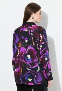 Kvinde med langt brunt hår iført en sort bluse med store lilla og røde blomstermønstre, vist fra ryggen mod en hvid baggrund.