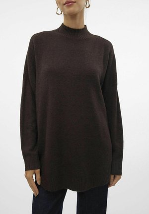 Femme portant un pull en maille marron foncé à manches longues avec poignets et col côtelés, associé à un jean bleu foncé et des boucles d'oreilles créoles dorées.