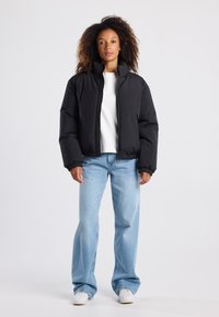 Giacca nera imbottita con chiusura a zip, colletto alto e polsini elasticizzati, indossata sopra una maglietta bianca e jeans a gamba larga blu.
