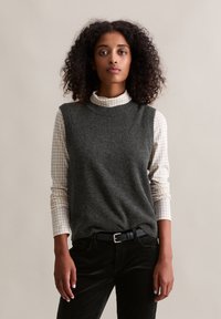 Grauer Wollpullover ohne Ärmel über einem langärmeligen, weiß-grau karierten Hemd, kombiniert mit schwarzen Cordhosen und einem schwarzen Gürtel.