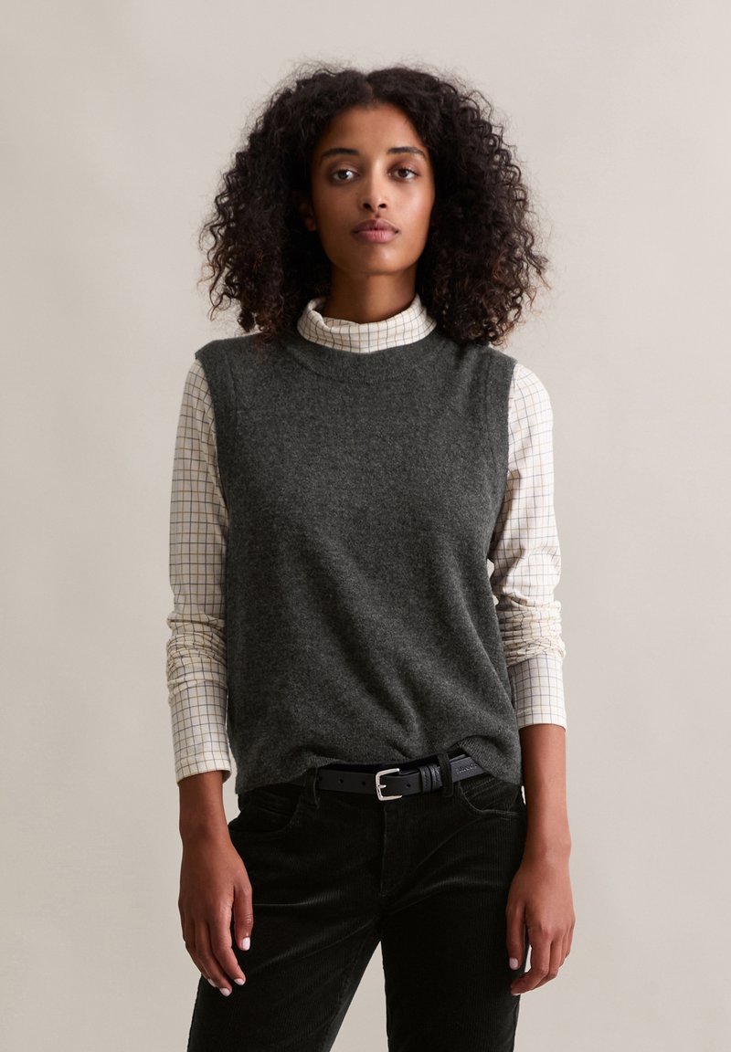 Grauer Wollpullover ohne Ärmel über einem langärmeligen, weiß-grau karierten Hemd, kombiniert mit schwarzen Cordhosen und einem schwarzen Gürtel.