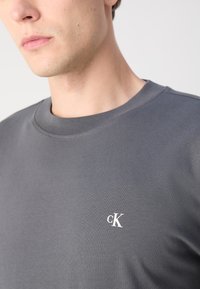 Šedé bavlněné tričko s kulatým výstřihem, hladkou texturou a malým bílým logem "cK" na levé části hrudi.