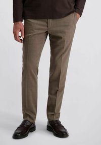 Man in een bruine pantalon en donkerbruin leren instappers, staand met één hand in de zak tegen een effen achtergrond.