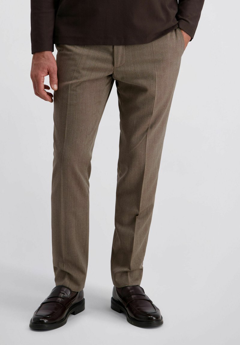 Man in een bruine pantalon en donkerbruin leren instappers, staand met één hand in de zak tegen een effen achtergrond.