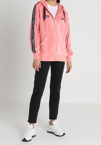 Sudadera rosa con cremallera en un material suave, que presenta una capucha, bolsillos frontales y una franja negra en las mangas con el logo. Combinada con pantalones negros y zapatillas blancas.