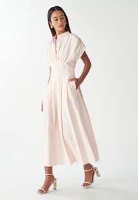 Vestido midi de cuello en rosa claro con rayas verticales blancas. Presenta mangas cortas abullonadas, una cintura ajustada y bolsillos laterales. Combinado con tacones blancos.