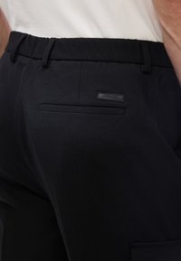 Κοντινό πλάνο μαύρου παντελονιού με πίσω τσέπη welt και ετικέτα λογοτύπου Calvin Klein στη ζώνη, σε συνδυασμό με λευκό πουκάμισο.