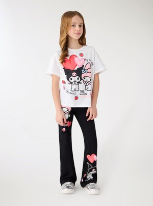 Ragazza giovane che indossa una maglietta bianca e pantaloni neri con personaggi dei cartoni animati, cuori e fragole, in piedi davanti a uno sfondo bianco.