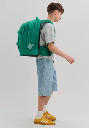 Boy wearing gray t-shirt, denim shorts, yellow sneakers, and a large green backpack with chest and waist straps walking right.