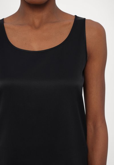 Donna che indossa un top senza maniche nero con scollatura a barchetta, che mostra il collo, le spalle e la parte superiore del torso su uno sfondo semplice.