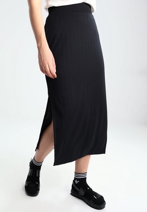 Mujer con falda midi negra acanalada con abertura lateral, calcetas negras con rayas blancas y sandalias negras de punta abierta sobre fondo blanco.