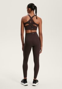 Braun Sport-BH mit ausschnittartigen Details und passenden hochgeschnittenen Leggings. Der Stoff ist glatt mit matter Oberfläche. Schwarze Sportschuhe.