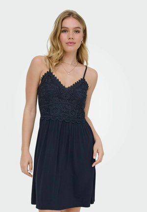 Jonge vrouw draagt een mouwloze zwarte jurk met een kanten lijfje en dunne bandjes, staand tegen een effen lichte achtergrond.