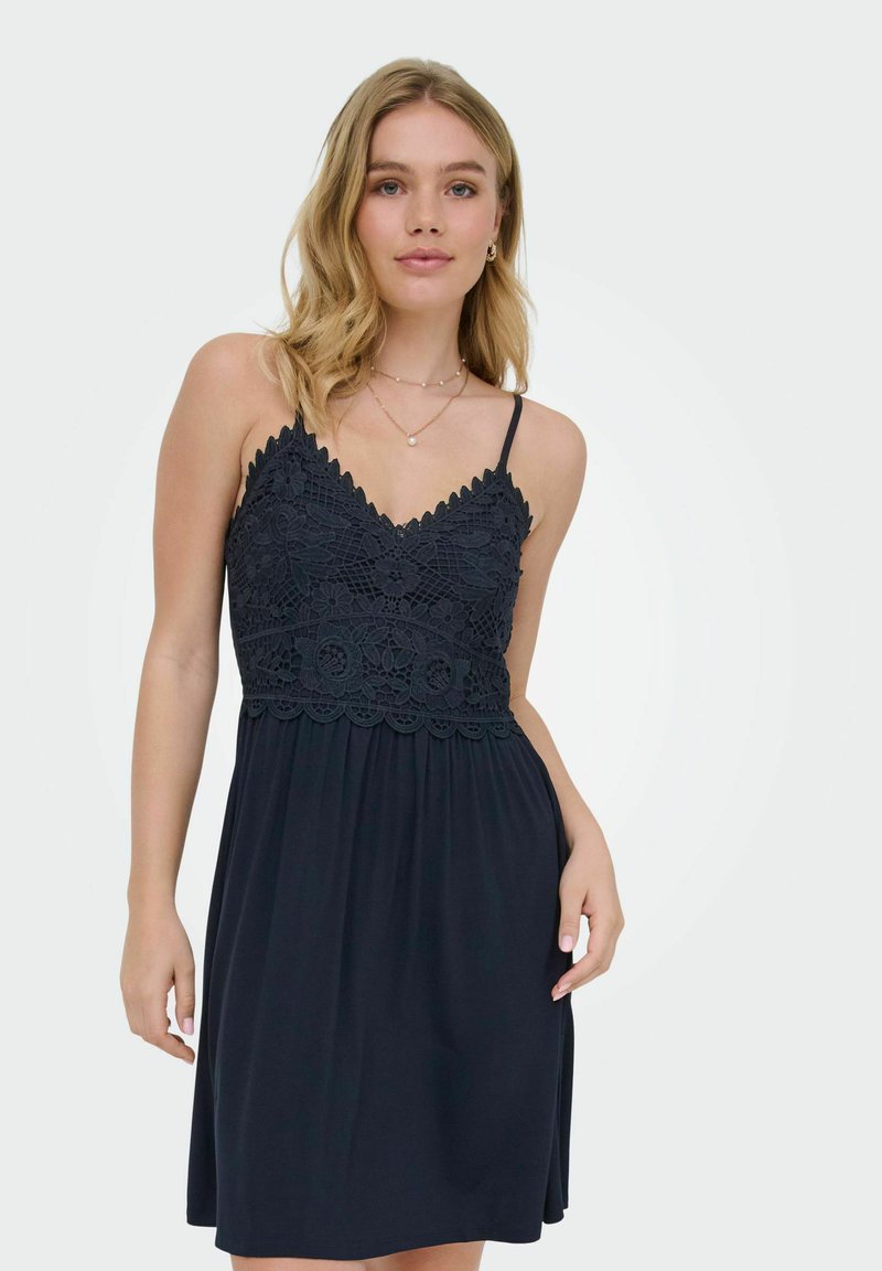 Jeune femme portant une robe noire sans manches avec un corsage en dentelle et des bretelles fines, debout devant un fond clair uni.