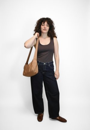 Débardeur marron, jean large en denim foncé, et chaussures marron à enfiler. Sac à main beige avec une seule bandoulière. Cheveux bouclés. Fond blanc uni.