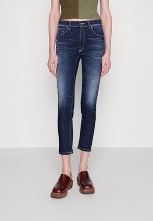 Jean slim bleu foncé avec détails délavés, porté avec des sabots en cuir marron et un débardeur court côtelé vert olive.