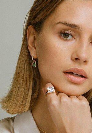 Goldring mit einem großen klaren Stein und feinen Details am Band; grüne Edelstein-Hoops; glatter, neutraler Hintergrund.